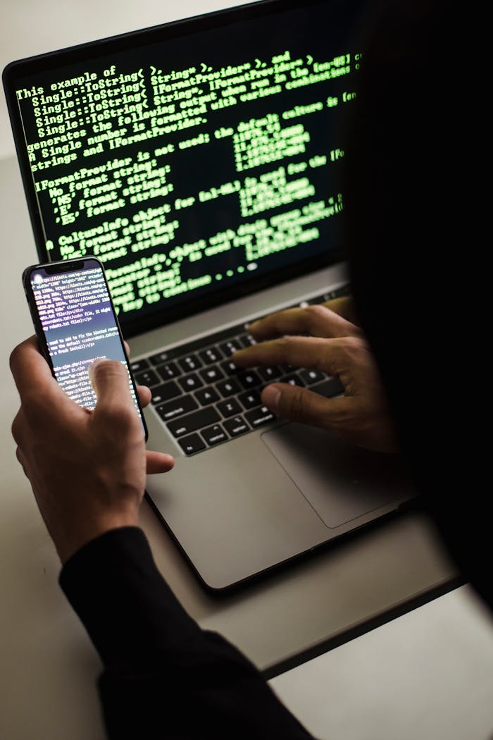 Home Unrecognizable hacker with smartphone typing on laptop at desk